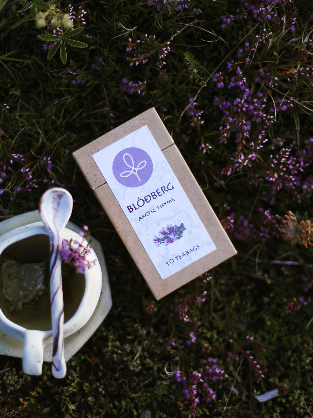 A kraft paper tea box labeled 'BLOBBERG Arctic Thyme' lies on a bed of green moss and small purple flowers, with a white ceramic teacup and spoon nearby.
