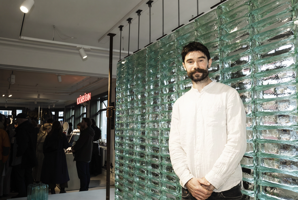 Man standing in a modern interior setting with glass wall and blurred people in the background