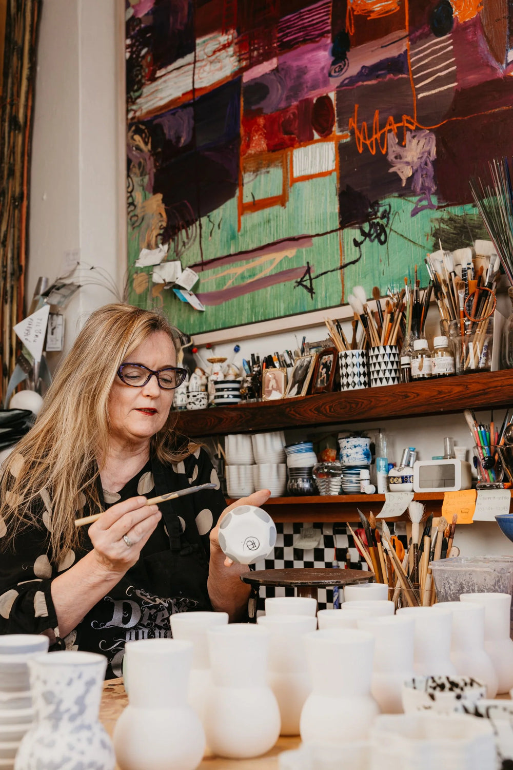 A woman with glasses and long blonde hair sits in an art studio, holding a white ceramic pot while painting. Shelves behind her are filled with paintbrushes, paint tubes, and art supplies, with a colorful abstract painting hanging on the wall.
